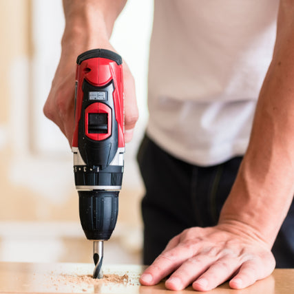 DIY RentalsA person in a white shirt uses a red and black power drill on a wooden surface, creating sawdust. The scene conveys focus and manual work.