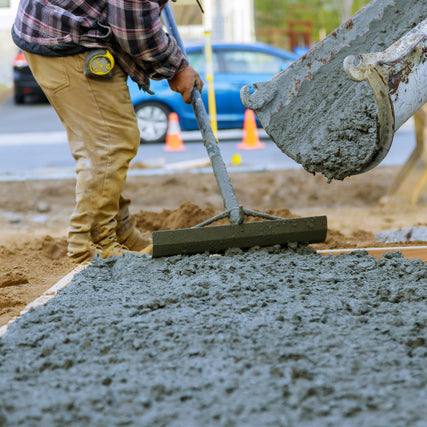 Contractor RentalsA worker in a plaid shirt and beige pants spreads wet concrete with a tool. A concrete mixer pours fresh mix. Traffic cones and a car are in the background.