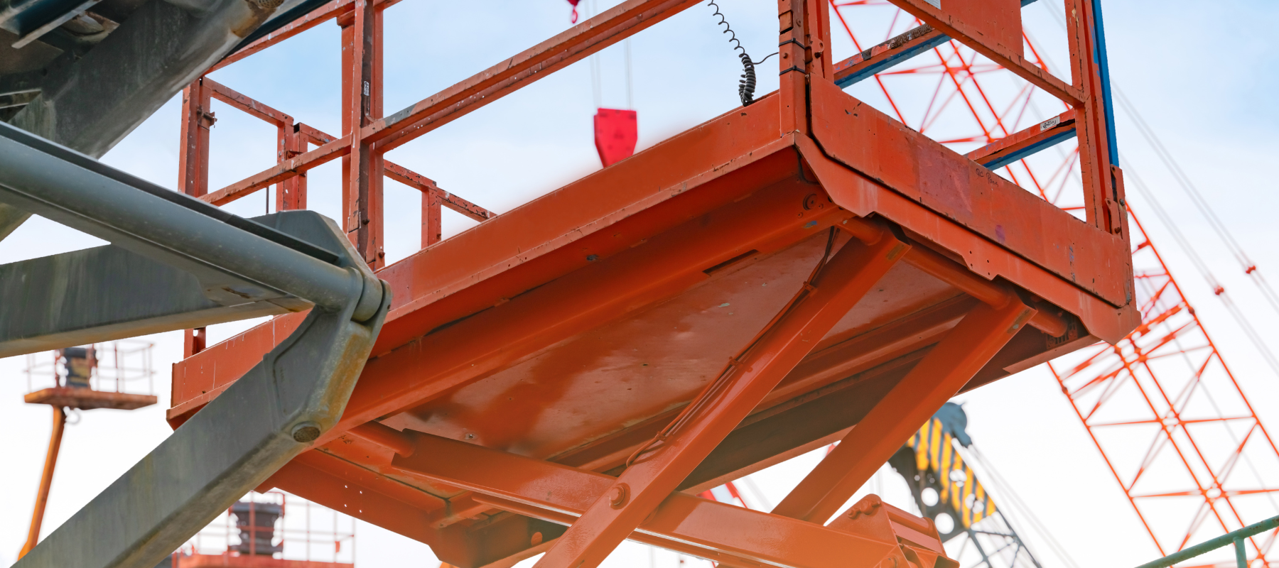 Orange scissor lift platform elevated against a backdrop of construction cranes and blue sky. The scene conveys industrial activity and height.